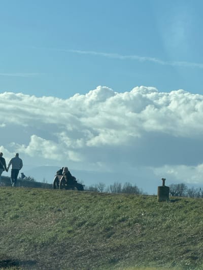 The image displays an outdoor scene in Altenheim, Germany, under a bright blue sky with extensive white cumulus cloud formations occupying the mid-section. A faint contrail is visible in the upper blue sky. The foreground and midground consist of a green, grassy slope with patches of brown. On the left, two individuals are walking from left to right, with their backs partially visible. The person on the left wears dark clothing and carries a shoulder bag. The person on the right wears a light-colored jacket and dark pants; their hands appear joined. Centrally, two individuals are seated on a dark wooden bench with large, circular side supports, facing away from the viewer and positioned on the crest of the grassy area. To the right, a weathered, cylindrical stone or concrete pillar, topped with a metal T-shaped fixture, is embedded in the grassy slope. A distant line of leafless trees and a hazy, undulating landscape form the background beneath the clouds.