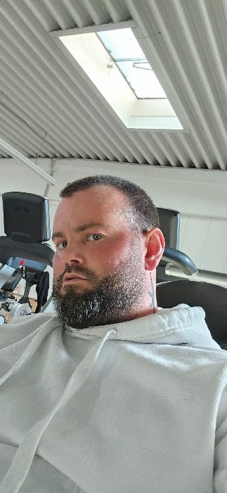 A male subject is depicted from the chest up, seated within an indoor fitness facility located in Rietberg, Germany. The individual has short, dark hair and a full, dark beard, with light-colored eyes directly engaging the camera. Visible redness is present on his cheeks. A dark, stylized tattoo is partially observable on the left side of his neck. He is wearing a light grey hooded sweatshirt with visible drawstrings. In the background, portions of dark exercise equipment are discernible. The overhead environment features a light-colored corrugated metal ceiling, containing a rectangular skylight that admits natural light.