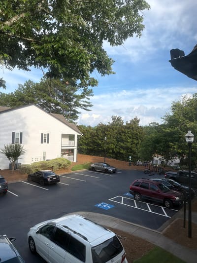 An exterior, high-angle, full shot captures a residential apartment complex parking lot under a partly cloudy sky. The apartment building is white with black shutters and balconies. Several cars are parked in the lot, including a black sedan, a silver sedan, a maroon SUV, and a white minivan. There are also multiple bicycles parked near a fence in the background. The scene is in Alpharetta, Georgia.