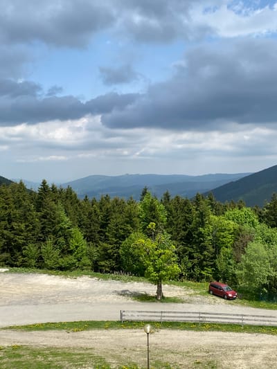 The image depicts a landscape with a view of mountains and trees, likely taken from an elevated position. In the foreground, there is a pathway or road running parallel to a line of trees. A red car is parked along the edge of the road, positioned near the trees. The road is marked with painted lines and a lamp post is visible on the side. The background features a mountain range under a sky with varying cloud cover, indicating changing weather conditions. The photograph appears to be taken at a location on the Mravenečník observation tower in Loučná nad Desnou, Czechia.