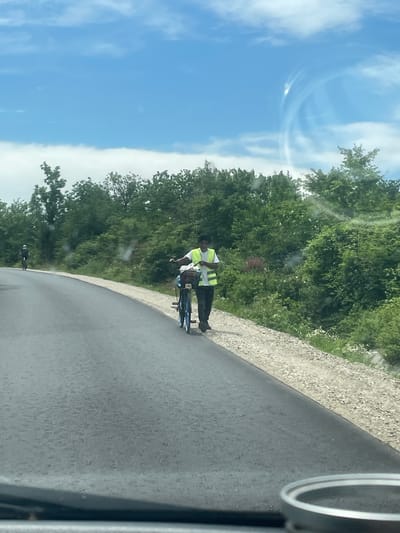 The image shows a man in a yellow vest standing next to a blue bicycle on a paved road, possibly on the outskirts of Probuda, Bulgaria. The road curves to the right, and on the left side of the frame is a line of trees and vegetation, followed by a blue sky with white clouds. On the right, there's a gravelly area next to the road. The photo appears to be taken from inside a moving vehicle.