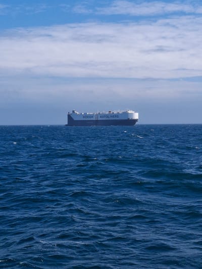 A large car carrier ship, identified by the text "HOEGH AUTOLINERS" on its side, is sailing on a dark blue, choppy sea under a bright blue sky with scattered white clouds. The ship is positioned in the center of the frame, viewed from a slightly low angle, giving it a dominant presence. The water exhibits numerous whitecaps, indicating moderate wave action. The horizon line is visible in the distance, separating the sea from the sky. The scene is devoid of any people or other vessels. The location is not specified in the image and cannot be determined.
