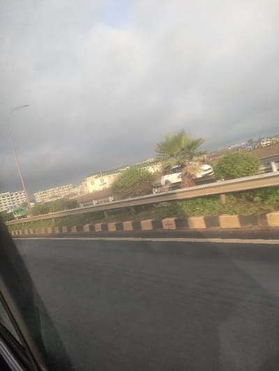 The image depicts a roadside scene in Theta ward, Kenya, captured from within a vehicle. In the foreground, a section of dark asphalt highway is visible, marked by a single white lane stripe. To the right of the roadway, a silver metal guardrail runs horizontally, supported by a segmented curb constructed from alternating light-colored and black rectangular blocks, one of which bears a faint red cross marking.

Behind this barrier, an upward-sloping, vegetated embankment features dense green shrubs and a prominent palm tree. A white passenger vehicle is observed partially embedded in this vegetation and the base of the palm tree, positioned behind the guardrail with its front obscured and its rear appearing elevated.

In the mid-ground, beyond the embankment, a row of light-colored buildings with distinct green sloped roofs is visible, one displaying the partial text "E.C. R-Bert." Further to the left, a large, multi-story building complex with numerous windows is present. To the far right, an elevated structure with some red components is partially visible.

The upper portion of the image is dominated by an overcast, predominantly grey sky. A tall, curved streetlight pole stands on the far left of the frame. No individuals are visible, and all depicted objects appear stationary.