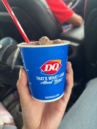 The image shows a person sitting in a car, holding a Dairy Queen (DQ) Blizzard. The cup is blue with the DQ logo and the slogan "THAT'S WHAT I LIKE About Texas." A red and white striped straw is inserted into the Blizzard. The person's hand is visible, holding the cup. The driver's seat and steering wheel are visible in the background, indicating the person is in a car. Based on the DQ brand and the "About Texas" slogan, the image likely originated in Texas, United States of America, but not specifically within Johnson City.