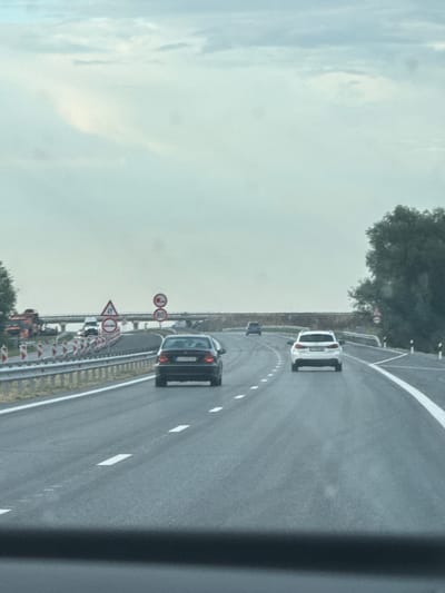 The image shows a view from a car traveling on a highway. The road has multiple lanes and is marked with white lines. Several cars are visible on the highway, including a black sedan in the foreground, a white SUV further ahead, and other vehicles in the distance. On the left side of the road, there are construction barriers and traffic signs, including a speed limit sign indicating 80. There is also a large orange vehicle, possibly construction equipment, on the left side. The sky is overcast with clouds. The scene is set on a highway near Bozduganovo, Bulgaria.
