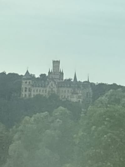 The image shows a pale, overcast sky above a hill covered in trees. Perched on the hill is a large, ornate building that appears to be a castle or a grand manor. The architecture is varied, with multiple turrets and gabled roofs. One prominent feature is a tall, square tower with smaller turrets. The building is light-colored, contrasting with the dark green foliage surrounding it. The overall scene is distant and somewhat hazy, suggesting it was photographed from a considerable distance or through some atmospheric obstruction. This building is Marienburg Castle, located in Nordstemmen, Germany.