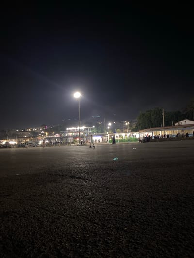 This is a nighttime, long-exposure photograph taken in Tipaza, Algeria, depicting a paved public space in front of a row of illuminated buildings. The foreground is dominated by a dark, textured paved surface, possibly asphalt, with patches of moisture reflecting the ambient light. In the mid-ground, a streetlamp with a bright, diffused light source stands prominently, casting a glow on the surrounding area. To the right of the streetlamp, a person is visible riding a kick scooter.

Further into the mid-ground, a line of buildings forms a backdrop. These structures are lit with various colors, suggesting they are shops, restaurants, or other commercial establishments. Many people are visible gathered in front of these buildings, some standing, others seated. They appear to be engaged in social activities or patronizing the establishments. The lighting creates a lively atmosphere.

In the background, more buildings and possibly housing are visible on a slope, also illuminated. Trees and vegetation can be seen interspersed with the structures. The overall scene suggests a popular gathering spot or promenade in Tipaza, illuminated for evening activity. The sky is dark, indicating the time of night.