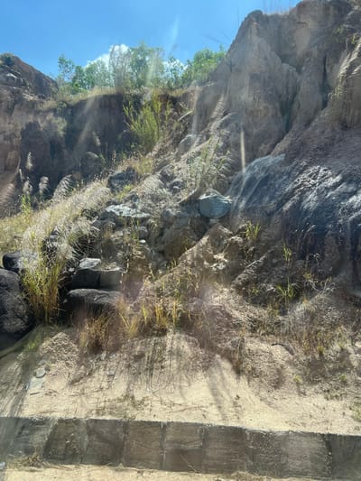A sunlit, rocky cliff face dominates the scene, partially obscured by dry grass and sparse green vegetation. Light filters through the vegetation, creating bright spots. The cliff descends to the right, meeting a sandy path bordered by blocky, concrete steps. Shadows are cast across the path, contrasting with the bright sunlight hitting the stone and sand. The sky appears blue in the background.