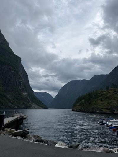 This is a wide, landscape photograph of the Gudvangen fjord in Gudvangen, Norway. The image is dominated by dark, imposing mountains that rise steeply on either side of a tranquil, dark body of water. The sky is overcast with heavy, grey clouds, creating a somber and dramatic atmosphere. In the foreground, a wooden pier juts out into the water, with a few small boats moored alongside it on the right. A red life preserver hangs on the pier. The shoreline on the left is a mix of rocks and a paved path. The overall impression is one of natural grandeur and a quiet, somewhat isolated, coastal setting.