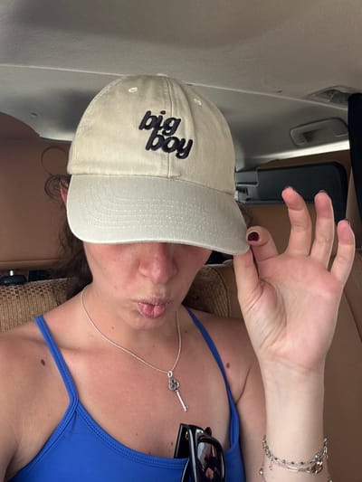 A woman in a bright blue swimsuit and a tan "big boy" baseball cap is in an unknown location in the United Arab Emirates. She is positioned within a vehicle; the interior walls and seats appear to be tan leather or a similar material. The woman is making an "okay" gesture with her hand. She has a silver necklace with a clover-shaped pendant and multiple bracelets on her wrist. Sunglasses are also visible resting on the swimsuit. Her fingernails are painted with a dark color.