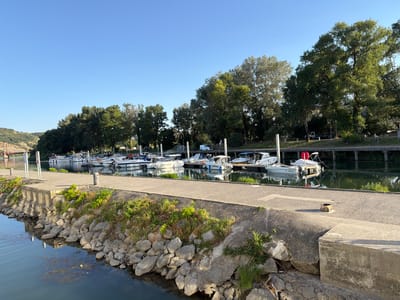 This image depicts a serene marina scene on a clear, sunny day, likely in the early morning or late afternoon given the long shadows cast. The foreground shows a stone embankment with various-sized rocks, some with sparse green vegetation growing between them, bordering a calm body of water that reflects the sky and surrounding trees. A concrete walkway runs along the top of the embankment, leading towards a dock lined with several small to medium-sized white motorboats.
The boats are moored to wooden posts and appear to be unoccupied. Some have white canopies, and one boat clearly displays a license plate with the text "ST 439354". Behind the boats, a line of mature trees forms a lush green backdrop, suggesting a natural or park-like setting. In the distance, beyond the trees, hints of buildings and a bridge can be seen, along with a hilly landscape under the bright blue sky. The overall atmosphere is peaceful and tranquil, characteristic of a quiet marina. The location is identified as Viviers, France, which fits the description of a picturesque waterside town. No people are visible in the frame, contributing to the stillness of the scene.