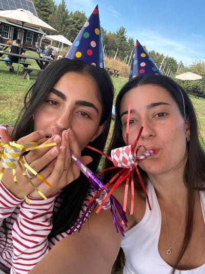 Two young adult females are depicted in a close-up, selfie-style photograph taken outdoors in Accord, United States. Both subjects wear dark blue party hats with multicolored polka dots. The female on the left, wearing a red and white striped long-sleeved shirt, holds a yellow and gold tinsel party blower with both hands near her mouth. A silver ring is visible on her left ring finger. The female on the right, in a white tank top and a silver necklace with a circular pendant, is actively blowing a red and purple tinsel party blower, with her lips pursed. A silver hoop earring is visible in her left ear. The background features a grassy outdoor area under a clear blue sky, with multiple wooden picnic tables, some shaded by white patio umbrellas. A building with a light-colored awning and solar panels on its roof is visible to the far left. Several individuals are seated at the picnic tables. Vineyard trellises and distant trees are present on the right, completing the rural outdoor setting.