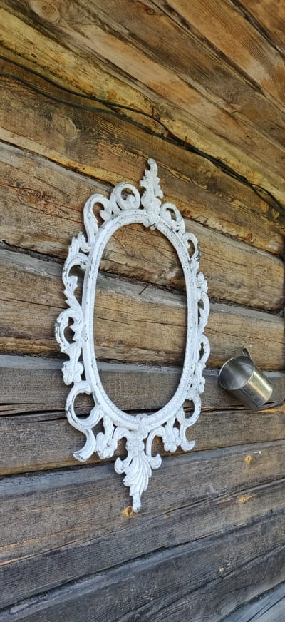 An ornate white oval picture frame hangs on a weathered wooden wall. To the right of the frame, a small silver metal cup is attached to the wall. The wall is constructed of horizontal wooden planks, some of which appear to be logs. The lighting suggests it is daytime, and the overall scene has a rustic aesthetic. The location is identified as Kalodzishchanski Sel'ski Savet, Belarus. There are no people visible in the image.