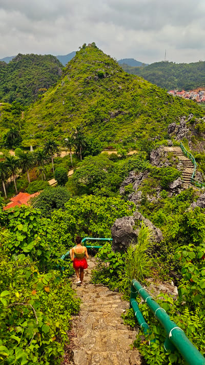 A woman with dark hair in a bun, wearing a yellow tank top and a red skirt, descends a stone staircase. She is walking away from the viewer, down a path surrounded by lush green foliage and rocky outcrops. The staircase is lined with a bright green metal railing.

The scene is a hilly, tropical landscape under a cloudy sky. In the background, more green, tree-covered mountains rise, with a small village of red-roofed houses nestled in the distance. The overall setting appears to be a park or tourist area, with winding paths and palm trees visible among the vegetation. The time of day is likely daytime, given the illumination, and the weather appears overcast. The environment is natural and verdant, suggesting a warm climate. The woman's posture and direction of movement indicate she is exploring or moving through the landscape.