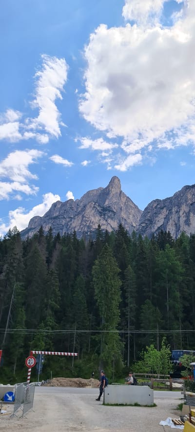 This is a vertical, eye-level outdoor shot taken on a sunny day in Prags - Braies, Italy. The image features a dramatic mountain landscape with a prominent, pointed peak dominating the upper left quadrant. The mountain is rocky and imposing, with visible striations and textures. Below the mountain, a dense forest of dark green evergreen trees forms a thick band across the mid-section of the image. The sky is a vibrant blue, scattered with fluffy white clouds, some of which are large and billowy while others are wispy.

In the lower right portion of the image, there is a glimpse of a paved area, possibly a road or pathway, bordered by temporary barriers and construction materials, including wooden pallets. Two individuals are visible near a concrete barrier. One, wearing a dark shirt and backpack, is leaning over the barrier, appearing to interact with something or someone on the other side. Another person, also with a backpack, is further back, looking away from the camera. A striped barrier arm, red and white, is positioned across the pathway, and a circular red sign with a white border and a "2" in the center is attached to it, likely indicating a height or weight limit. A blue directional arrow sign is also visible on the right side of the frame.

The overall scene suggests a natural, mountainous environment with some signs of human activity or development. The lighting indicates it is daytime, likely late morning or early afternoon, given the brightness of the sky and the shadows. The presence of the barriers and construction materials hints at ongoing work or preparations for an event.