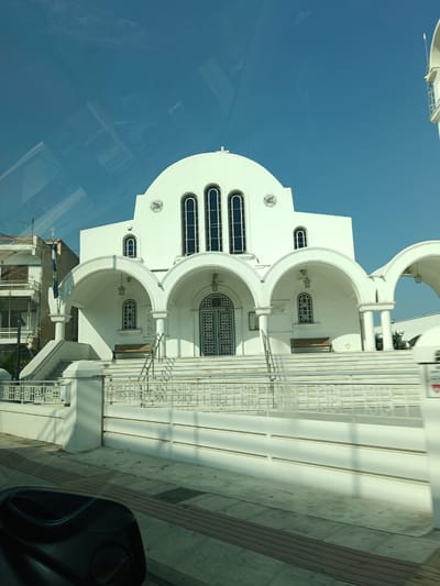 The image shows the exterior of a white, domed church with arched porticos and classical architectural elements. The church has tall, arched windows with dark frames and decorative metal grilles on some of the lower windows. A set of wide, white stairs leads up to the main entrance, with white railings on either side. There are benches on either side of the entrance. To the left of the church, a multi-story building with balconies is visible. A Greek flag is flying from a flagpole. The sky is clear and blue. The foreground shows a sidewalk and the edge of what appears to be a car's interior, suggesting the photo was taken from a vehicle. The specific location is the Municipal Unit of Velos, Greece, and the church is likely the Church of St. Nicholas in Velos.