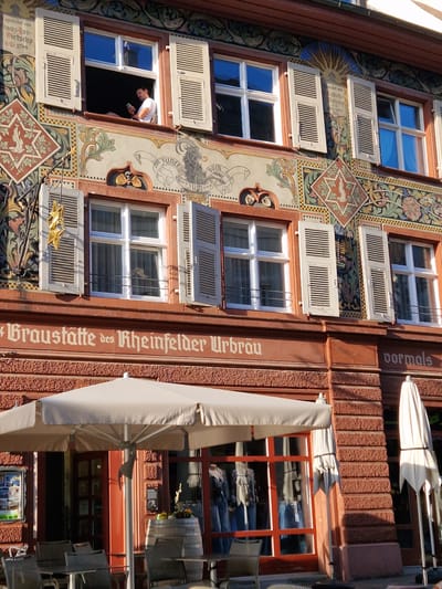 Here's a detailed description of the image:

**Scene:** The image depicts the facade of a building in Rheinfelden, Switzerland.

**Architecture:** The building features a traditional architectural style, possibly a historic structure. The facade is decorated with painted designs and patterns. The building has multiple windows with white shutters. The building has an inscription that reads "Braustätte des Rheinfelder Urbräu vormals". This translates to "Brewery of the Rheinfelden Urbräu formerly". There is a decorative, possibly golden, ornamental piece sticking out the building.

**People:** A man is leaning out of an open window and looking down. He is wearing a white shirt and is holding what appears to be a cell phone.

**Objects:** There are umbrellas and outdoor seating, suggesting a restaurant or bar.