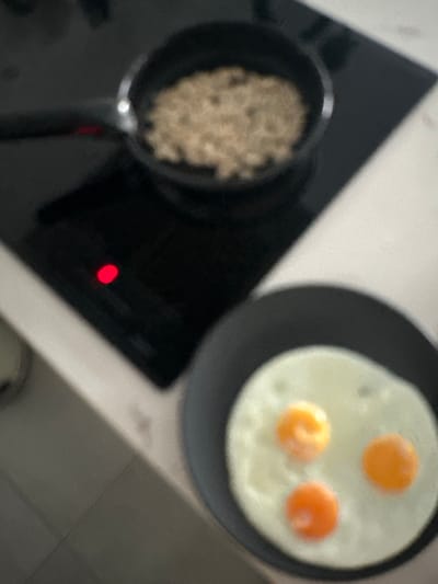 The image shows a close-up, slightly blurry view of a kitchen counter in Vilanova i la Geltrú, Spain. On a black induction hob, a red indicator light is illuminated. To the right of the hob, a black frying pan contains three fried eggs with visible yolks. Behind the frying pan and slightly to the left, another black pan is on the hob, containing a light-brown, granular substance, possibly oatmeal. The kitchen counter appears to be white.