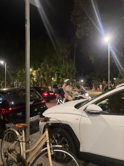 This nighttime image captures an urban street scene in Sestri Levante, Italy, adjacent to a public park or green space. The scene is illuminated by multiple bright streetlights, casting long light rays.
In the immediate foreground, a light-colored (beige/cream) bicycle with a brown seat and handlebars is parked vertically, leaning against a prominent metal lamppost that bisects the left side of the frame. To the right of this lamppost, the front passenger side and partial hood of a white SUV are visible, appearing to be parked. To the left of the lamppost, the rear of a black Hyundai i20 is parked, displaying an Italian license plate "M 620PR".
In the mid-ground, a large, light-colored motorcycle is in motion, moving from left to right. Two individuals are on the motorcycle: the driver, wearing a helmet and light-colored clothing, and a passenger behind, partially obscured. Behind the parked black Hyundai, a dark-colored sedan with illuminated red taillights is also in motion.
The background consists of a densely vegetated area, consistent with a park, featuring numerous dark trees, including palm-like varieties. Several indistinct human figures are visible within this park area, particularly on the right, some appearing near what may be playground equipment. The ground visible is paved, indicating a street or promenade.
