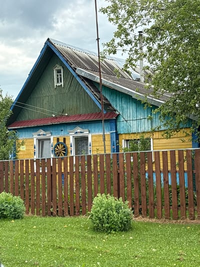 A traditional wooden house, painted green on the upper gable and blue and yellow on the lower sections, stands behind a brown wooden fence. The house has a corrugated roof and a small attic window on the gable. Two windows are visible on the main floor, each adorned with white carved trim, and a decorative wooden wheel hangs between them. The yard is covered in green grass with two bushy green plants in the foreground. The sky is overcast. The scene appears to be in a rural setting.