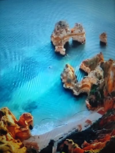 An aerial view showcases a coastline featuring rocky formations in the turquoise sea. A prominent rock arch and isolated stack are visible. The coastline displays eroded cliffs with patches of vegetation, contrasting with the sandy beach below. The water appears clear, revealing varying depths around the rock formations. The scene is bathed in sunlight, highlighting the texture of the rocks and the vibrancy of the water.
