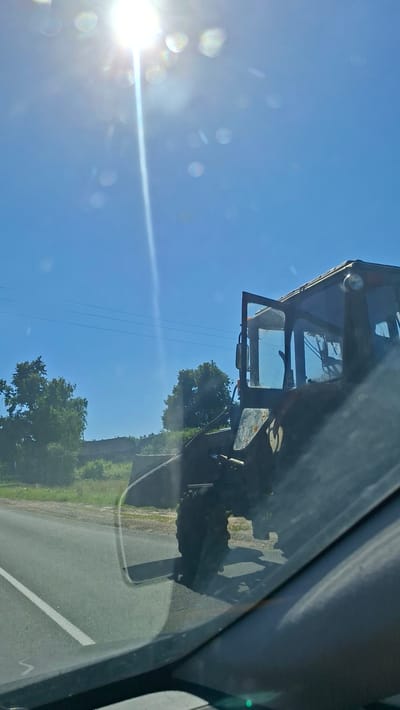 A dark-colored tractor is parked on the side of a paved road, likely in the vicinity of Хажоўскі сельскі Савет, Belarus, under a bright, sunny sky with visible sun flare and lens artifacts. The tractor's cab door is open, and its large rear tire is prominent. Green trees and foliage are visible behind the tractor, and a few power lines are strung across the sky in the distance. The foreground shows a portion of a car's interior, including the windshield, suggesting the image was taken from inside a vehicle.