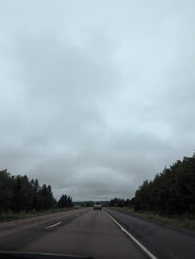 A shot from inside a vehicle on a highway in Steeves Mountain, Canada, shows a gray, overcast sky with fluffy clouds. A line of dark green trees runs along the sides of the road, with more trees visible in the distance. A silver pickup truck drives away from the camera on the right lane of the highway. The road is asphalt with white lines marking the lanes.