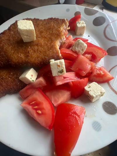 The image shows a plate of food, likely in a domestic setting, as indicated by the dishware. The food consists of two breaded cutlets, possibly chicken or pork, served with chopped tomatoes and cubes of cheese. Some of the cheese cubes appear to have herbs mixed in. The plate has a speckled pattern with brown and pinkish-brown circles. The lighting suggests it might be daytime, possibly near a window, given the reflections on the plate. There is no indication of the specific location within Augsburg, Germany, or any people present. The focus is solely on the food presentation on the plate.