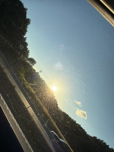 The image captures a sunlit scene, possibly from inside a vehicle, looking out through a window with visible dust or water spots. The perspective is angled, with the foreground dominated by a dark, possibly upholstered surface on the right and a diagonal line suggestive of a window frame or dashboard. A road, marked by white lines, runs diagonally across the lower left portion of the frame, bordered by dense, dark green foliage that appears to be trees or bushes. In the upper right, a clear blue sky is visible, with a few small white clouds and a bright, blurred sun. The sun is low in the sky, casting a warm glow and creating lens flare. There is a hint of a vehicle's reflection or interior in the very bottom center of the image. The scene appears to be a roadside view, potentially along a highway, outside of Stenungsund, Sweden, given the typical landscaping and road infrastructure found in such areas.