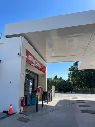 The image shows the exterior of a "Smartshop" convenience store located in the city of Komotini, Greece. The store is situated under a modern white canopy, likely part of a gas station. The facade of the shop is white with a prominent red sign that reads "Smartshop" and smaller text below it saying "eat drink shop". To the left of the entrance, there is a white wall with a red fire extinguisher mounted on it, and an orange traffic cone. Inside the shop, a Red Bull refrigerator and a display of drinks are visible. Several black bollards are positioned in front of the shop. The ground in front of the store is paved with concrete, and there are several rectangular grates. The background features lush green trees and a clear blue sky. The scene appears to be peaceful and undisturbed.