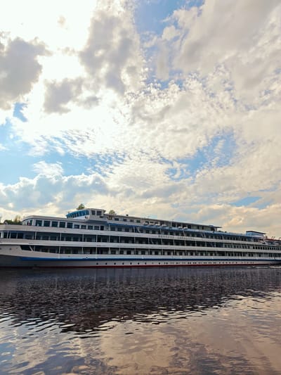 The image shows a large white cruise ship on the water. The ship has multiple decks with numerous windows. A blue stripe runs along the lower part of the ship's hull, with a thin red stripe beneath it. The water reflects the ship and the sky. The sky is filled with white clouds and some blue sky. The location is Водник Снт, Russia.