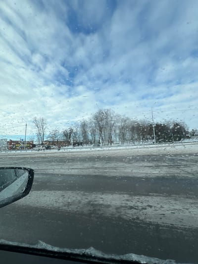 The image depicts a winter street scene in Sambek, Russia, viewed from inside a vehicle. The foreground shows the lower-left section of a vehicle's windshield and a side mirror, both partially obscured by water droplets and ice accumulation along the bottom edge of the windshield. The midground features a multi-lane asphalt road, partially covered with patches of snow and ice, indicating wintry conditions. To the left of the road, a commercial area is visible with several low-rise buildings, some displaying colored signage, and a parking lot containing multiple parked automobiles. A streetlight post stands on the left. To the right of the road and extending towards the center, a dense line of mostly bare deciduous trees and some evergreens suggests a park or wooded area. Another streetlight post and overhead power lines are visible on the right. The background consists of a sky with scattered white and light-grey clouds against a blue backdrop, also viewed through the droplet-covered windshield. No individuals are visible.