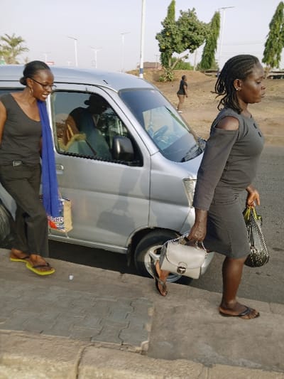 The image depicts an outdoor scene in Mumbwa, Zambia, featuring two adult females in the foreground alongside a silver minivan.
On the left, an adult female with dark skin and braided hair, wearing eyeglasses, a dark sleeveless top, dark trousers, and yellow flip-flops, stands facing partially towards the minivan. A blue fabric is draped over her left shoulder, and she holds a patterned bag. Her mouth is open.
On the right, another adult female with dark skin and braided hair, wearing a dark long-sleeved top with shoulder cut-outs, a dark knee-length skirt, and dark flip-flops, is depicted in motion. She is stepping onto or off a concrete curb, carrying a light grey shoulder bag with a metallic clasp in her right hand and a larger, darker patterned bag in her left hand.
A silver minivan is positioned between the two women. A blurred reflection of a person, possibly wearing a hat or cap, is visible in the driver's side window. The immediate foreground consists of grey interlocking concrete pavers and a concrete curb. Beyond the curb, a darker paved surface abuts the minivan. The background features an unpaved, light-colored area where a distant figure in dark clothing stands. Several trees, including a round-canopied tree and two tall, slender trees, are visible in the mid-background. Streetlights or utility poles are also present. The sky is clear and light, indicating daytime, with lighting suggestive of late afternoon or early morning.