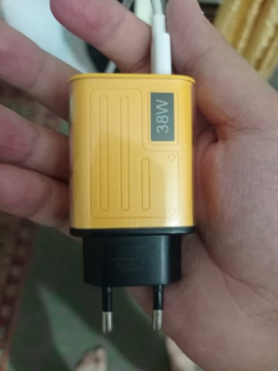 A person's hand holds a yellow and black USB charger with "38W" printed in white text on a gray rectangular background on the yellow part of the charger. The charger has two metal prongs extending from the black part. The background is blurred and appears to be indoors with hints of carpet and a curtain. The image does not provide any information or context related to the city of Chust, Uzbekistan.