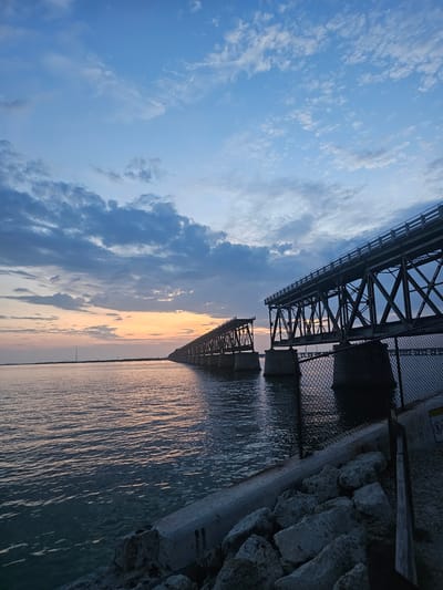 The image shows a view in Big Pine Key, United States, featuring a bridge over water at sunset. The sky has a mix of light blue and gray clouds, with the setting sun casting an orange and golden glow on the water's surface. The bridge is constructed of metal and has a mesh fence. A concrete path with a rock barrier runs along the edge of the water.