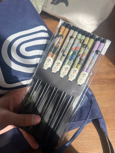 A person's left hand is centered, holding a clear plastic-wrapped package containing five pairs of decorative chopsticks. Each chopstick handle features a unique color scheme and traditional Japanese-style female figure, while the lower sections are black. The package is backed by black cardboard. To the left and beneath the package, a dark blue fabric bag with a white arched logo is partially visible. The background consists of a wooden surface and a blurry white object with an indistinct dark silhouette. Strong light illuminates the scene from the upper left. The location is the Hof Ashkelon Regional Council, Israel.