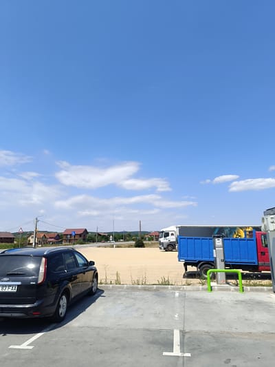 In Poietari, Romania, on a clear, sunny day, a black Ford station wagon is parked in a concrete lot. In the background, a large blue truck is parked next to a red truck. The blue truck has a graphic of a lion's head on its side. A green metal structure, likely a tire inflator, is situated in front of the trucks. Beyond the trucks, houses with red roofs are visible, and further in the distance, trees and rolling hills can be seen. The sky is a vibrant blue with scattered white clouds.