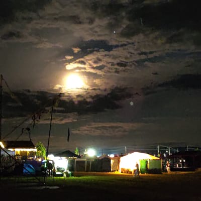 The image depicts a nighttime scene in Kudrino, Russia, likely a market or festival. The sky is dark with scattered clouds illuminated by a bright full moon, casting a glow on the surroundings. Below, several tents and stalls are visible, with some lit up, suggesting activity. In the foreground, a grassy field is present. There are a few blurry figures of people walking or standing near the tents. Structures, possibly temporary buildings or booths, are set up. The overall impression is that of an outdoor event taking place at night, with artificial lighting from the tents contrasting with the natural moonlight.
