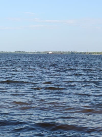 A wide shot shows a calm river under a clear blue sky with a few wispy clouds. In the distance, a dense line of green trees forms the opposite bank. Two tall, slender industrial chimneys are visible on the distant shore, one with red and white markings. A large, white barge with a dark hull is moving across the water in the middle distance. The foreground is dominated by the rippling surface of the river, reflecting the light and creating a textured pattern of blues and grays.
