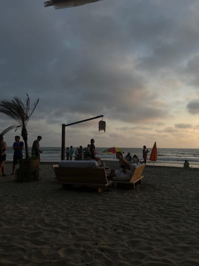 The image captures a beach scene in Montañita, Ecuador, at coordinates -1.821 latitude, -80.756 longitude. The scene is bathed in the soft light of dusk, with a partly cloudy sky above. In the foreground, there are two wooden benches arranged in a semi-circular shape on the sand, accommodating a group of individuals. One woman is seated on the rightmost bench, facing the camera. She wears a white tank top and a black bathing suit bottom. Several other individuals are in the background, standing around the benches, engaged in conversation. Among them, one woman is wearing a white sundress. A few other individuals are standing further back, some on the beach, and others partially obscured by a palm tree. A large, partially obscured palm tree is situated to the left of the benches, adding a tropical touch to the scene. The benches are positioned close to the shoreline, with the ocean waves gently rolling in the background. A vibrant yellow beach umbrella provides a pop of color near the water's edge. An orange beach umbrella is also visible in the background, adding a second splash of color to the scene. A light fixture attached to a pole, resembling a chandelier made of seashells, is positioned behind the benches. This fixture is slightly obscured by branches of the palm tree. The overall atmosphere is relaxed and peaceful, depicting an evening scene on a beautiful tropical beach. The sand, although not clearly defined, appears smooth and light. The scene captures a moment of leisure and enjoyment on a tranquil beach.