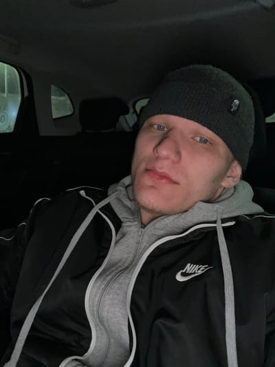 A male individual is depicted in a close-up, head-and-torso shot, seated inside a vehicle in Dobryanka, Russia. The man has light skin, blue eyes, and visible stubble on his jawline and chin. He wears a dark gray knitted beanie, a light gray zippered hoodie, and a black jacket with white piping and a white Nike logo on the right chest. He is looking directly at the camera with a neutral expression. The vehicle interior is dark, with elements of the ceiling and seat headrests visible in the background. Through the left-side windows, faint ambient light illuminates indistinct exterior structures, including a white surface with a circular or "0" marking. The scene appears to be documented at night or in low-light conditions.