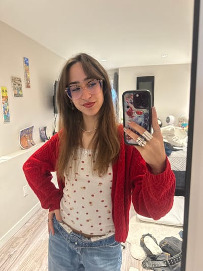 A woman poses for a selfie in a mirror. She wears light-wash jeans, a white top with red dots, and a red cardigan. Her hair is brown and she sports glasses with pink frames. Several large silver rings adorn her fingers. The phone case is decorated with a colorful design. The background shows a room with artwork on the wall and a bed with a white comforter.