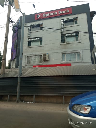 The image shows the facade of a building in Sokuluk, Kyrgyzstan, housing "Optima Bank." The building is a multi-story structure with a light-colored, corrugated metal exterior. The top of the building features a prominent red sign with the bank's name and logo. There are several windows on the building, some of which are covered with metal grates. An outdoor air conditioning unit is visible on the right side of the building. A large purple advertisement is displayed on the left side of the building, featuring text and what appears to be a QR code. Electrical wires and poles are visible in the foreground and attached to the building. A corrugated metal fence or barrier runs along the lower portion of the building. In the bottom right of the frame, the front corner of a blue car is visible, and a timestamp reads "19.06.2026 11:52". The environment appears to be an urban or semi-urban setting with a cloudy sky.