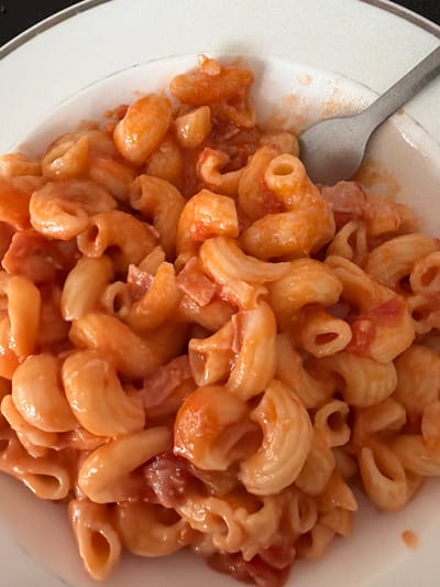 The image depicts a close-up of a bowl of pasta, specifically elbow macaroni, coated in a reddish-orange sauce. A metal spoon is partially visible on the left side of the frame, positioned within the bowl. The pasta is piled high in the white bowl, with the focus primarily on the texture and color of the dish. The overall lighting is bright, accentuating the sheen of the sauce. There is no indication of location in Pentre, United Kingdom, or any people present.
