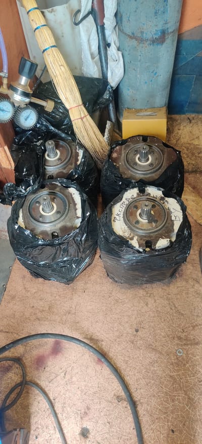 The image shows four components, possibly hydraulic pumps or motors, wrapped in black plastic bags. These components have a metal splined shaft protruding from a circular flange with several bolts. One of the components has some faded, handwritten text on its white surface, which appears to say "CK.ITPA.89".
In the foreground, there is a broom made of dried grass, secured with red and blue bands. To the left of the broom, there is a brass pressure regulator with two gauges. The gauges have white faces with black markings and needles. A purple hose is attached to the regulator.
The background includes a long, blue, cylindrical object with some rust and chipped paint, and a small yellow box. A black cable with some white lettering snakes across the brown, textured surface on the right side of the image. The overall scene appears to be an outdoor or workshop setting, with a slightly cluttered and used appearance. There are no people visible, and the lighting suggests it could be daytime, but there are no clear indicators of weather or a specific time of day. The location context provided is Rizunenkove, Ukraine.