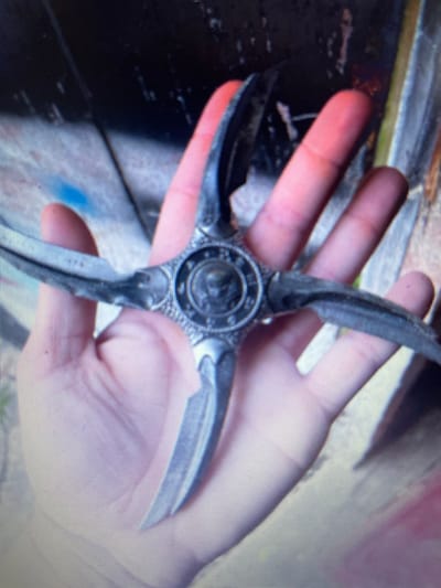 The image displays a human hand holding a multi-bladed metallic object. The scene is located in Noida, India.
The object is a silver-grey, four-bladed device with a central circular medallion. This medallion features an embossed skull design with dark accents, encompassed by a textured, patterned ring. Four distinct, sharp-edged blades extend radially from the central piece, each exhibiting a slightly curved profile. The metallic surface shows minor scuffs and variations in tone.
The hand, belonging to an individual with a light skin tone, is positioned palm-up, with fingers slightly spread, securely gripping the central portion of the object. The fingertips, particularly the index and middle fingers, exhibit a reddish hue.
The background consists of an indistinct, dark, and textured surface, possibly a wall, exhibiting irregular lighter streaks and a prominent blue mark visible towards the upper left portion of the frame. The overall lighting appears diffuse.
