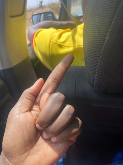 The image captures the interior of a vehicle in Unguwar Gaya, Nigeria, during daylight. In the foreground, a dark-skinned human left hand is prominent, with the index finger extended vertically upwards while the other fingers are curled into a fist. Direct sunlight illuminates the palm side of the hand and the extended finger. To the right, a dark, textured fabric surface, consistent with a vehicle seat or headrest, is partially visible. In the background, a person wearing a bright yellow garment with a red band on the upper arm is seated, their arm bent at the elbow. Through a vehicle window, a bright sky, distant sparse vegetation, and a portion of a side-view mirror are discernible.