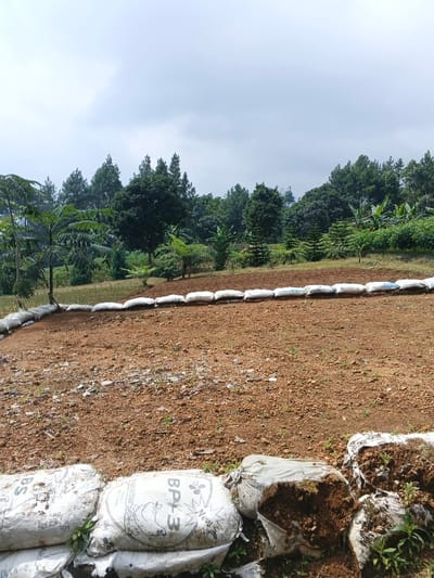 A plot of red-brown soil is surrounded by white sandbags. The bags form a low retaining wall, preventing soil erosion. In the background, a line of trees and vegetation extends across the horizon under a cloudy sky. The trees are a mix of pines and leafy species. The scene appears to be an agricultural project in a rural or hilly area.
