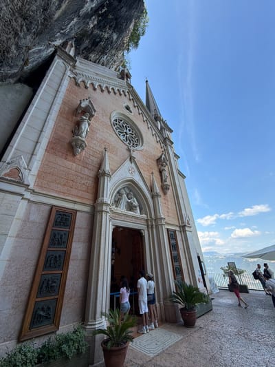 The image captures a side view of a church built into the side of a rocky cliff face, likely the Santuario della Madonna della Corona in Ferrara di Monte Baldo, Italy. The church has a light pink facade with decorative white trim and a large rose window above the entrance. Two statues are mounted on the facade, one on each side of the rose window. The entrance is an arched doorway with a relief sculpture above it, and a group of three young people are standing in front of it, looking inside. To the left of the entrance, there are four tall, narrow wooden panels with carved reliefs. Potted plants are placed on the paved area in front of the church. In the background, a scenic view of a valley with hills and a body of water under a blue sky with scattered clouds is visible. Several other people are seen walking or standing on a paved area to the right of the church, near an umbrella. The rocky cliff overhangs the top left of the church.