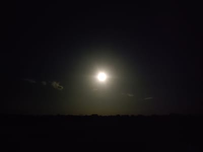 The image depicts a dark, nighttime scene with a bright, luminous object in the sky, likely the moon, which is surrounded by a halo of light. There are a few faint, wispy clouds in the upper portion of the sky. Below the sky, a dark, silhouetted line suggests the horizon, potentially the edge of a forest or a line of trees. The foreground is almost entirely black, obscuring any details on the ground. The location is specified as Cherevky, Ukraine. There are no people or discernible objects in the foreground or middle ground. The scene is still and quiet, with the dominant feature being the bright celestial body.
